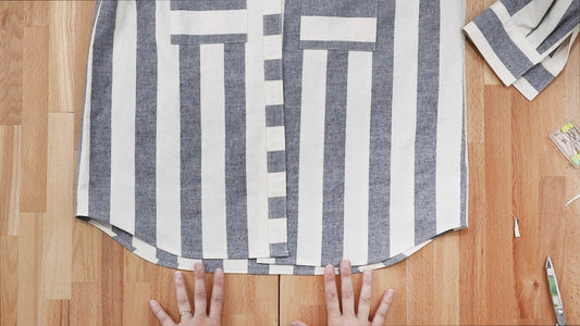 Archer Button Up in gray and white stripes laid flat on wood table, hands smoothing hem during hemming step of sew-along