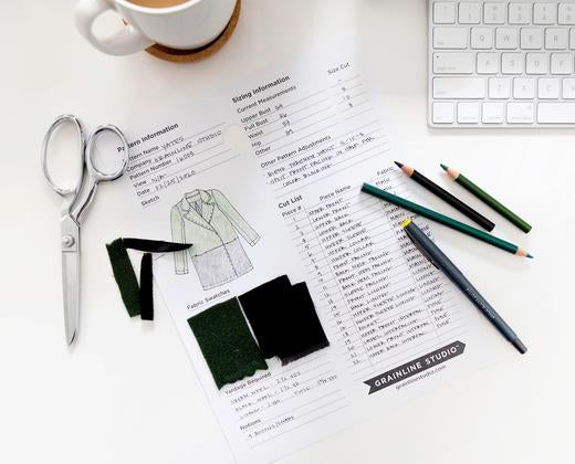 Grainline Studio pattern planning sheet flat lay with dressmaking shears, fabric swatches, colored pencils, mug, and keyboard