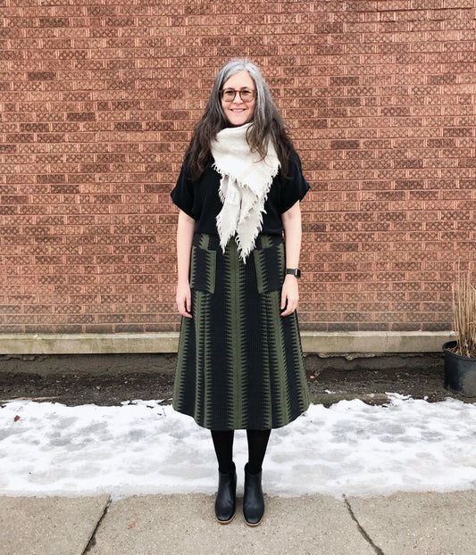 Reed Skirt in olive and black jacquard, worn with black tee, cream scarf, and black ankle boots against brick wall