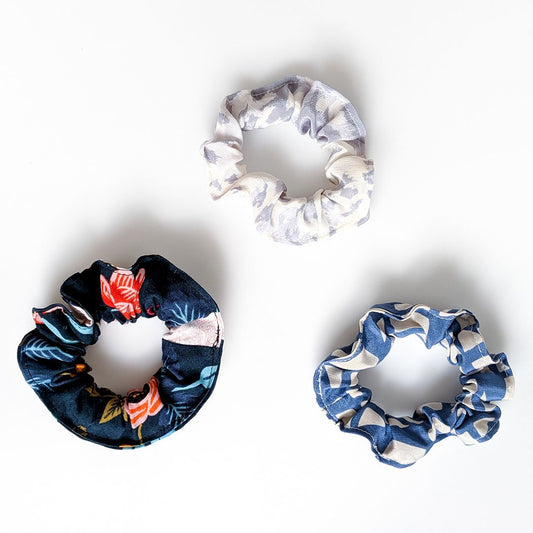 Three handmade fabric scrunchies on white background in gray animal print, navy tropical, and blue geometric prints