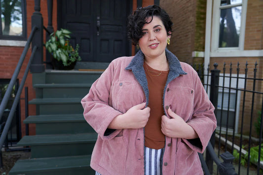 Size 28 model in pink corduroy Thayer Jacket with gray sherpa collar, rust Hemlock Tee, and striped Reed Skirt on city steps
