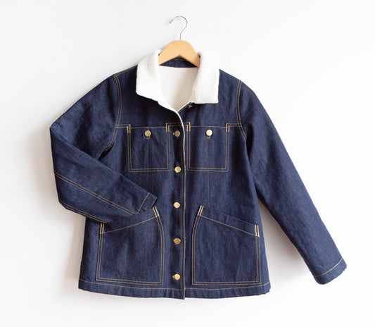 Thayer Jacket in dark denim with cream sherpa collar, gold topstitching, brass buttons, and patch pockets, on wooden hanger