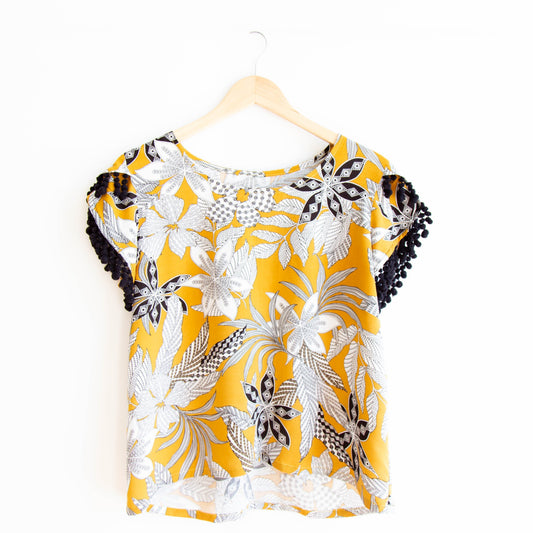 Scout Tee with petal sleeves in yellow, white, and black floral print with black pom-pom trim on sleeves, on wooden hanger