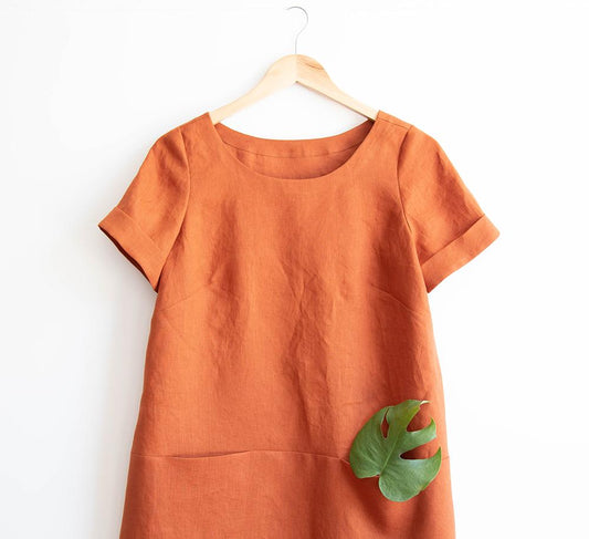 Uniform Tunic in rust linen with short cuffed sleeves and inseam pockets, monstera leaf tucked in one, on wooden hanger