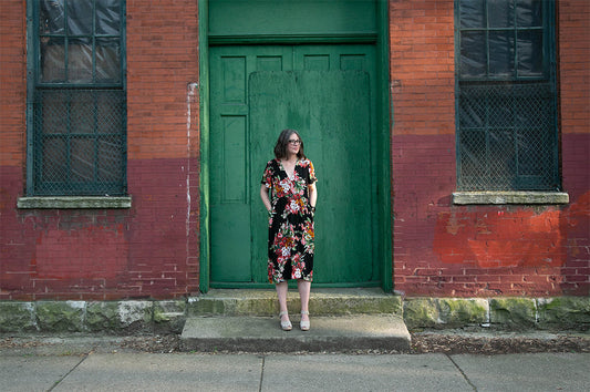 Woman wearing Felix Dress in black rayon with multicolor floral print, in front of weathered brick building with green door