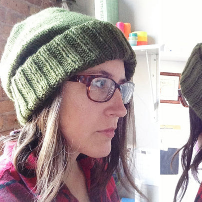 Person wearing a ribbed knit slouchy hat in army green with folded brim, glasses, and a red plaid shirt