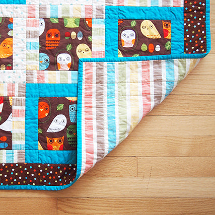 Owl baby quilt with patchwork blocks featuring brown owl print, turquoise binding, and striped backing on wood floor