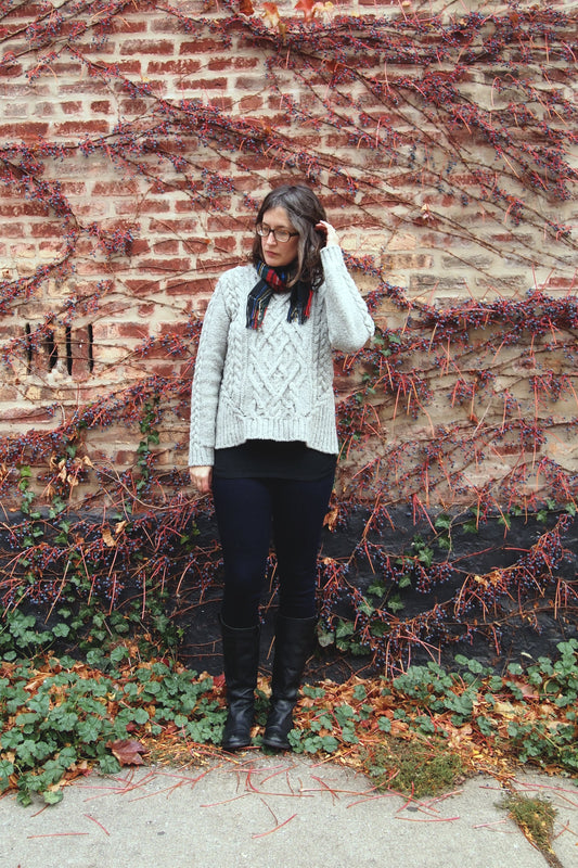 Person wearing Stonehenge cable knit sweater by Michele Wang with plaid scarf against berry vine covered brick wall