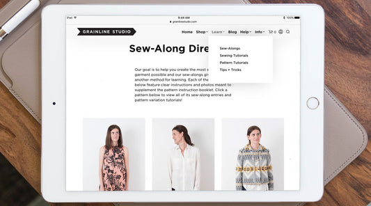 iPad showing Grainline Studio Sew-Along Directory with Learn menu open and three modeled garments on wooden surface
