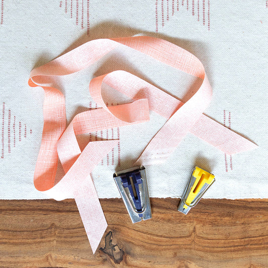 Pink crosshatch bias tape strips on cream linen with two bias tape makers in blue and yellow, on wood surface
