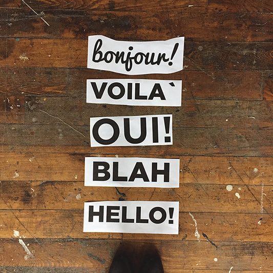 White text labels reading Bonjour, Voila, Oui, Blah, and Hello on weathered wood showing Linden Sweatshirt appliqué options