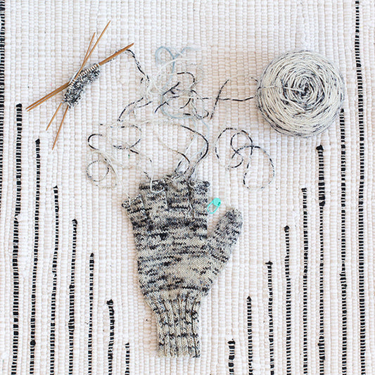 Knitting flat lay on a woven rug showing a gray marl fingerless glove in progress, with yarn, needles, and stitch markers