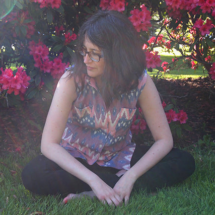 Woman sitting cross-legged on grass in a sleeveless Archer Button-Up in multicolor ikat print, pink rhododendrons behind her