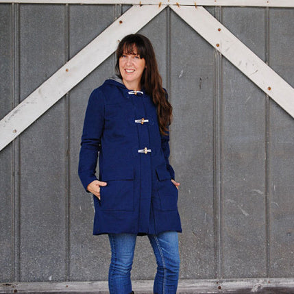 Person in navy Cascade Duffle Coat with toggle closures and patch pockets, worn over jeans against a corrugated metal wall