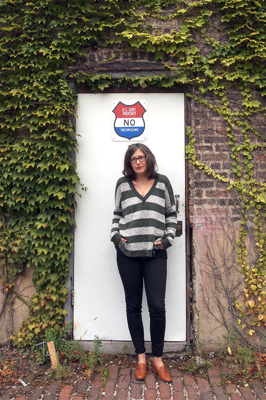 Person wearing green and gray striped V-neck sweater with black pants in front of ivy-covered door with no trespassing sign