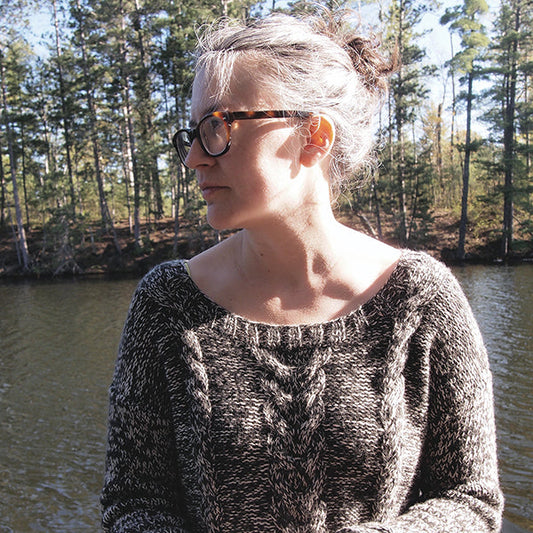 Person wearing Stone Lake Sweater in charcoal marl with cable knit detail, standing by lake with pine forest background