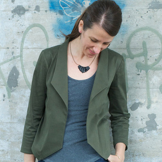 Woman wearing olive green Morris Blazer in stretch cotton twill over gray tank top, against textured concrete wall with graffiti