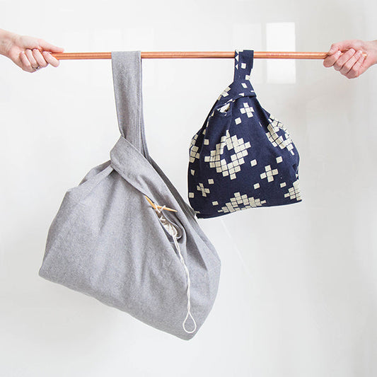 Two Stowe Bags hanging from a copper rod: light gray with knitting needles and yarn, and navy with cream diamond print