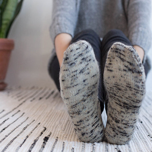 Close-up of handknit gray marl socks shown from the sole, worn on a woven rug with a gray sweater in the background