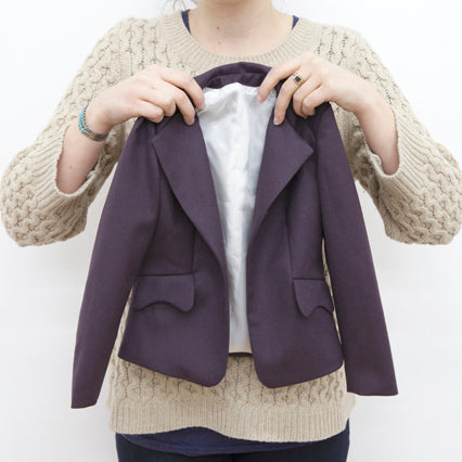 Person holding a mini purple blazer with notch lapels and white lining in front of a cream cable knit sweater