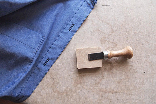 Blue chambray Archer Button-Up with finished buttonholes, laid flat next to a wooden buttonhole chisel and block