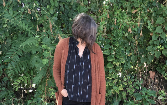 Person wearing rust cardigan over black ikat-print sleeveless top, standing in front of green foliage