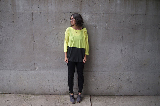 Person in a neon yellow and charcoal colorblock Hemlock Tee with three-quarter sleeves and black jeans against concrete wall