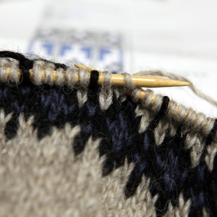 Close-up of knitting needle working stranded colorwork with black and tan yarn showing Fair Isle technique in progress