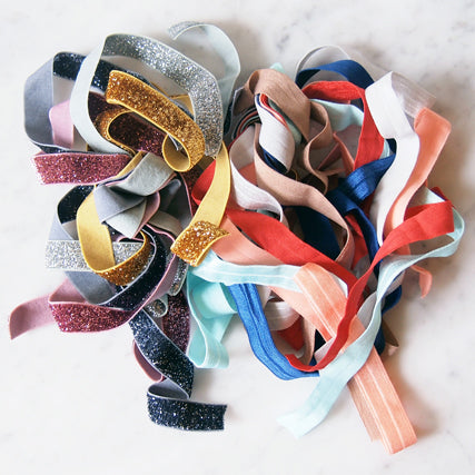 Fold-over elastic in various widths including glitter, velvet, and solid styles in gold, pink, navy, red, and coral on marble