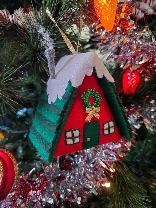Handmade felt cabin ornament with red walls, green striped roof, snow trim, and gold wreath, hanging on a decorated tree