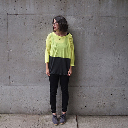 Person in a neon yellow and charcoal colorblock Hemlock Tee with three-quarter sleeves and black jeans against concrete wall
