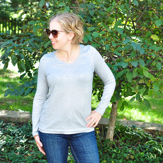 Woman wearing long-sleeved Lark Tee in light gray jersey with V-neck, styled with jeans and sunglasses in an outdoor setting