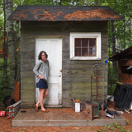 Woman in a gray top and denim Moss Skirt standing in the doorway of a weathered wooden shed in a wooded setting