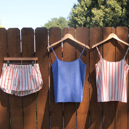 Three handmade pajama pieces hanging on a wooden fence: red striped shorts, solid blue camisole, and red striped camisole