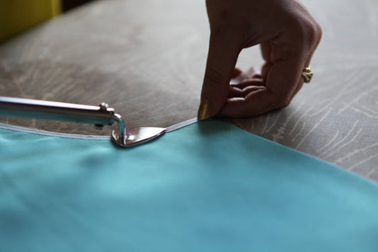Hands pressing white lightweight interfacing to delicate teal silk chiffon fabric neckline on ironing board