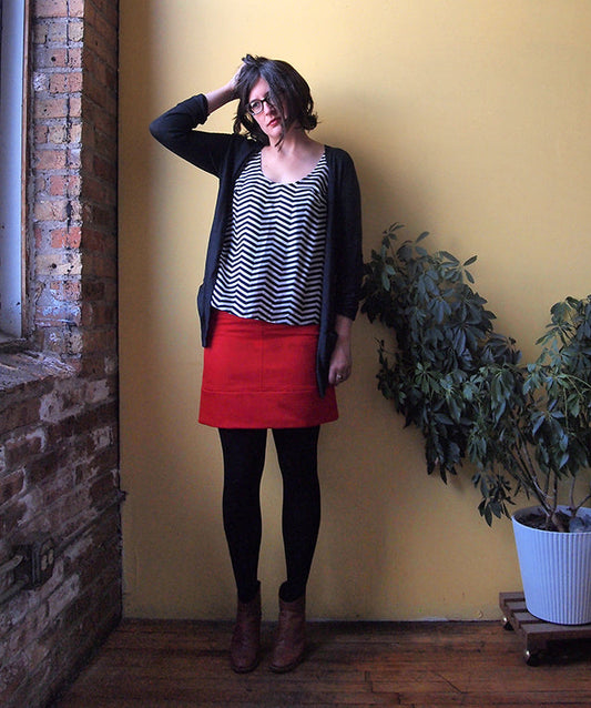 Woman in black cardigan, chevron tank, red wool Moss Skirt, black tights, and brown boots against a yellow wall with brick