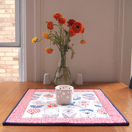 Quilted placemat with pink gingham border and floral center, with a white ceramic candle holder and orange ranunculus flowers