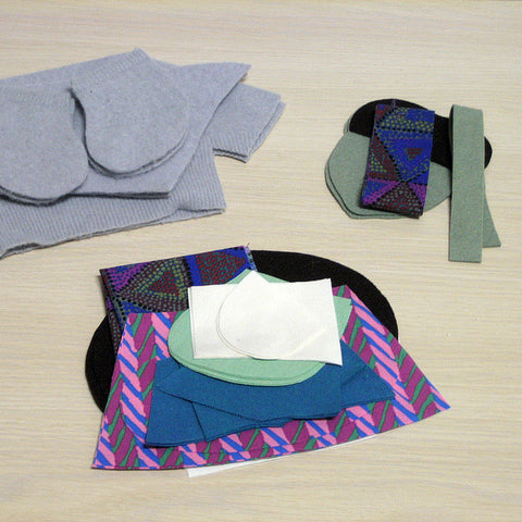 Cut fabric pieces for doll clothing including dusty blue felt sweater, geometric print dress, and felt acorn backpack