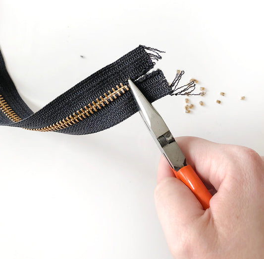 Hand using orange-handled pliers to clamp a new zipper stop onto black zipper tape, with removed brass teeth scattered nearby