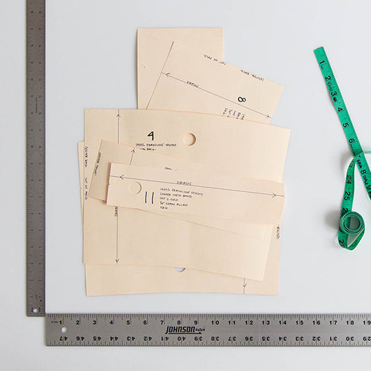 Oaktag pattern pieces with handwritten notes, two rulers forming a right angle, and green measuring tape on white surface