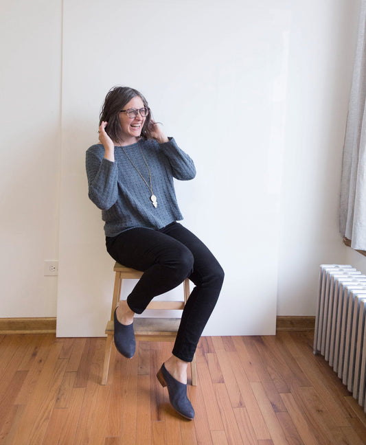 Person sitting on wooden stool wearing blue handknit pullover, black jeans, and pendant necklace, smiling and adjusting hair
