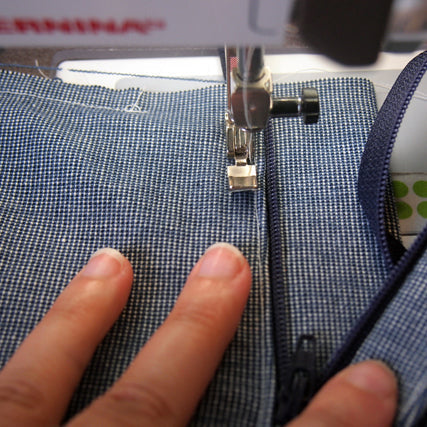 Hands guiding blue textured fabric under a sewing machine presser foot while inserting a fly front zipper