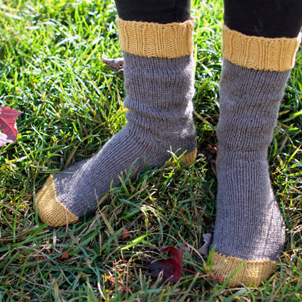 Hand-knit socks in heather gray with mustard yellow ribbed cuffs and heels, worn outdoors on grass with fallen autumn leaves