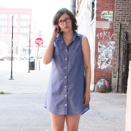 Woman wearing a sleeveless lavender blue chambray button-up shirt dress on a city street with a brick wall background