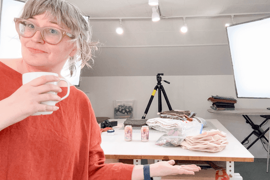 Jen Beeman in red dress holding mug at filming worktable with fabric samples, light stand, camera, and tripod in studio