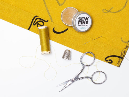 Hand sewing flat lay with yellow thread, embroidery scissors, thimble, needles, and Sew Fine Thread Gloss on mustard fabric