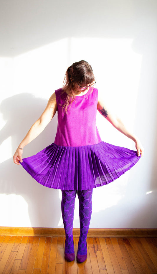 Person wearing Uniform Tunic in magenta sleeveless top with purple pleated skirt and patterned tights, holding out the skirt