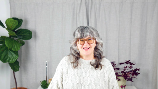 Jen Beeman smiling in light gray handknit cable sweater and rose glasses, with fiddle leaf fig and oxalis plants