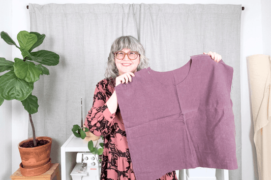 Jen Beeman in pink floral Poppy Dress holding unfinished purple linen Bernina Top, with plants and serger in background