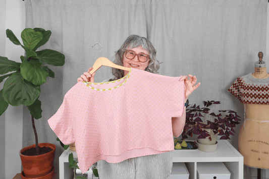 Jen Beeman holding finished Bernina Top in pink swiss dot with yellow gingham neck binding on hanger, plants and dress form